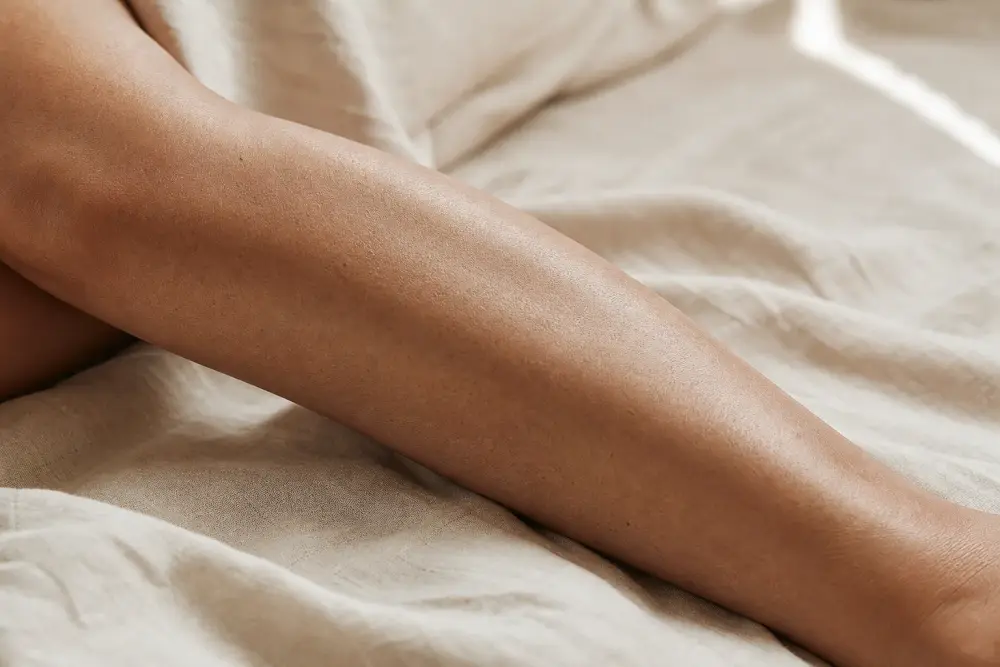 Jambe posée sur un tissu clair, texture de peau naturelle, lumière douce