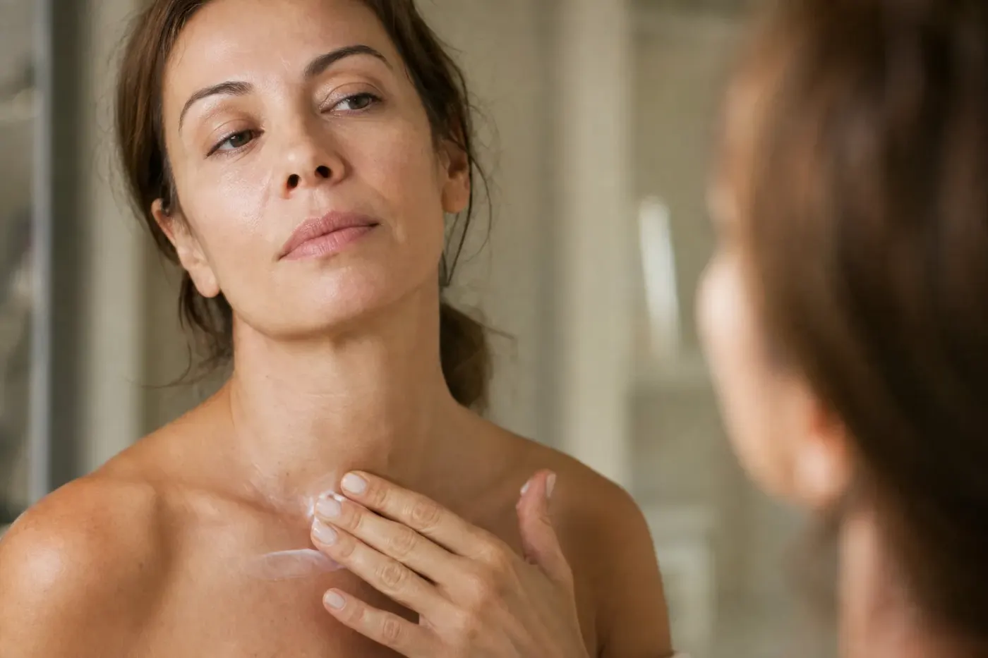 Femme appliquant une crème du bout des doigts sur son cou et son décolleté devant un miroir