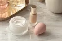 Pot de crème ouvert, flacon de fond de teint et éponge rosée sur une coiffeuse claire