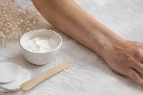 Bol de crème blanche, spatule et coton près d’un avant-bras posé sur une serviette claire