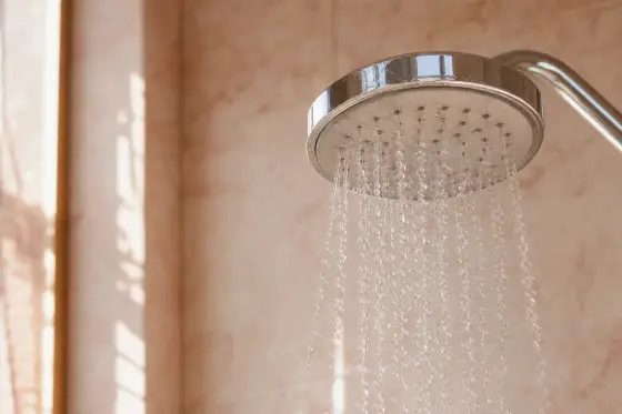 Eau claire s’écoulant d’un pommeau de douche chromé devant un mur carrelé clair, en lumière naturelle