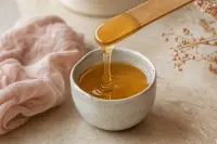 Pot de cire tiède et spatule en bois soulevant un ruban de cire ambrée sur un plan de pierre claire