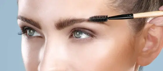 Main tenant une pince à épiler près d’un sourcil brossé, devant un miroir