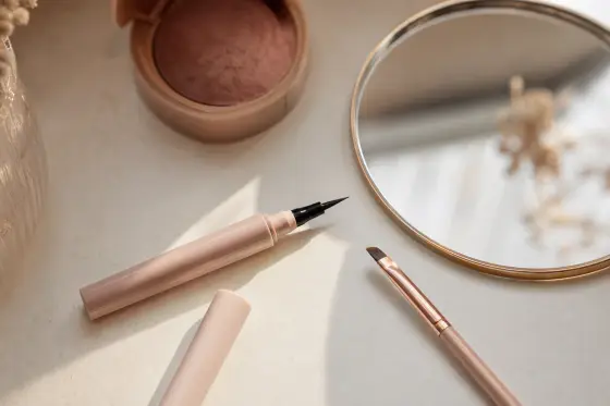 Eyeliner liquide noir ouvert, pinceau biseauté et petit miroir sur une coiffeuse claire