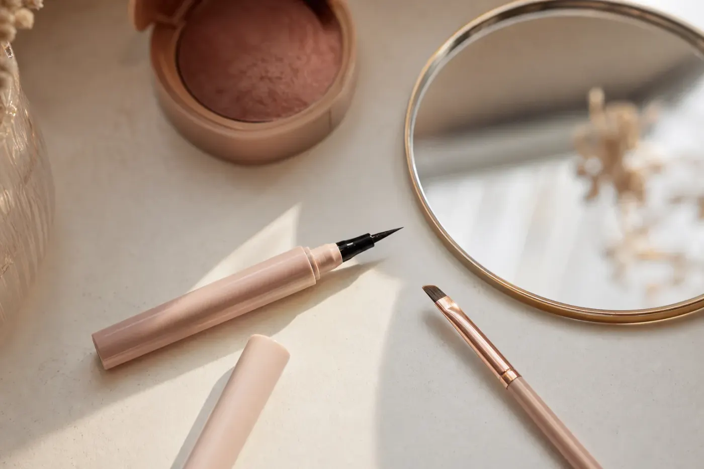 Eyeliner liquide noir ouvert, pinceau biseauté et petit miroir sur une coiffeuse claire